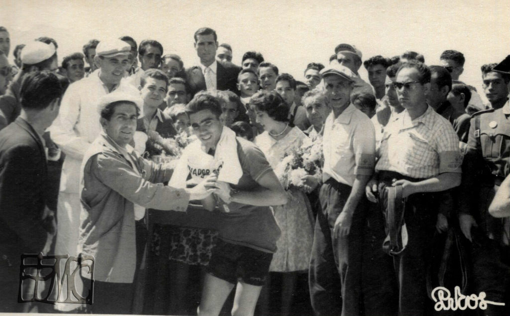 Salvador Honrubia recibe el maillot de ganador de la 3ª etapa en 1959 ayudado por Doroteo Cabañas. Detrás pueden verse a Paco Pin (mono blanco y gorra) la madrina Pili Muñoz, Casimiro Rubio y Basilio Delgado con el brazalete de comisario.